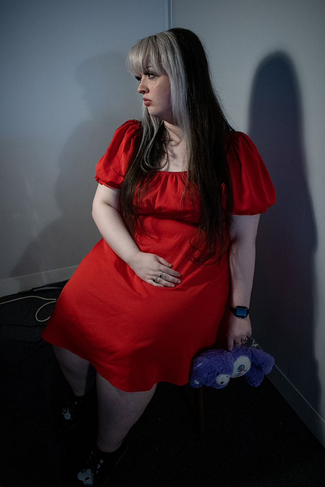 Amy Hannah: Epitaph | Thursday 22 June  – Wednesday 19 July 2023 | Atypical Gallery | Image: photo of a figure in a corner; almost-waist-length black and grey hair, head turned away from the camera; wearing a strong-red, unpatterned, knee-length dress; holding a blue-and-white teddybear in left hand 