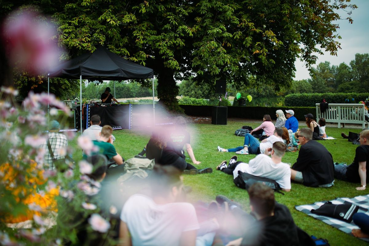 Emerging Patterns / Homebeat, IMMA Outdoors 2021 / Photo: Molly Keane | IMMA Outdoors | June – September 2023  June – September 2023 | IMMA | Image: Emerging Patterns / Homebeat, IMMA Outdoors 2021 / Photo: Molly Keane  – people sitting on the grass in IMMA grounds, looking towards what appears to be a DJ performing 