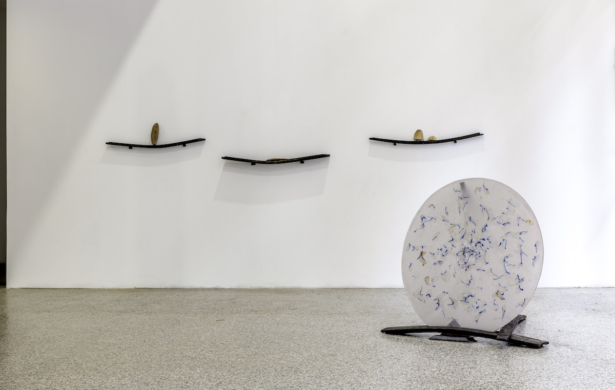 Rosie O'Reilly: shifting silence, cuttlefishbone, plant epoxy & woad dye, 2022; image credit Louis Haugh | Athrú, an Invocation | Friday 7 July  – Sunday 3 September 2023 | Luan Gallery | Image: Rosie O'Reilly: shifting silence, cuttlefishbone, plant epoxy & woad dye, 2022; image credit Louis Haugh – in the right foreground is a translucent white disk tilted almost vertical and held in place by what appear to be black wooden planks of the type you could imagine being salvaged from a shipwreck; three similar planks are attached to the the wall behind in the exhibition space, and each appear to have one or more cuttlefish bones on them, propped against the wall or lying flat 