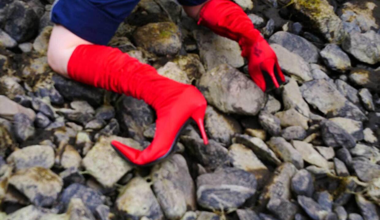 Uprooting Tallaght – Performance Art Festival | Tuesday 1 August  – Saturday 5 August 2023 | Rua Red | Image: photo which seems to show the arm and leg of a person, or a person (we see only the arms, or one arm and one leg, or two legs) wearing a crimson high-heeled, knee-high boot on one limb, an identically coloured elbow-length glove on the other; this is on a surface that looks like rock on a shore 