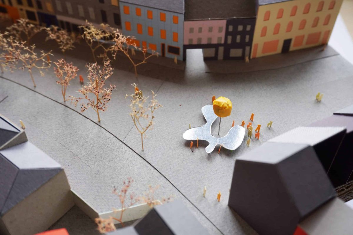 A model of Urban Mirror, Plattenbaustudio’s installation on Cornmarket Street | Island City: Commission | deadline Tuesday 1 August 2023  | National Sculpture Factory | Image: A model of Urban Mirror, Plattenbaustudio’s installation on Cornmarket Street – We see a 3D model of a street, seen from above and to the side; a few buildings at a three-way intersection; small, flat people in orange or cream; a sculptural installation consisting of a flat, horizontal, organic, metal shape vaguely resembling a Miró figure or an amoeba; where the metal shape curves in deeply, there is a pole, maybe 4 metres high (to scale), with an orange ball on top, which is maybe 2 metres in diameter  