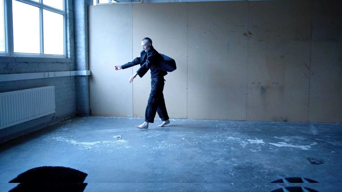 Sandra Johnston: An Instinct, A Continuance | Saturday 22 July  – Saturday 26 August 2023 | GOMA Gallery of Modern Art | Image: probably a still from a video – we see the artist in what may be a roomy studio space with a large window in the wall to the left, a concrete floor and an MDF (?) wall behind; there is something, a sort of chunky flour, on the floor; the artist is in motion towards the window wall, with arms outstretched downward gaze