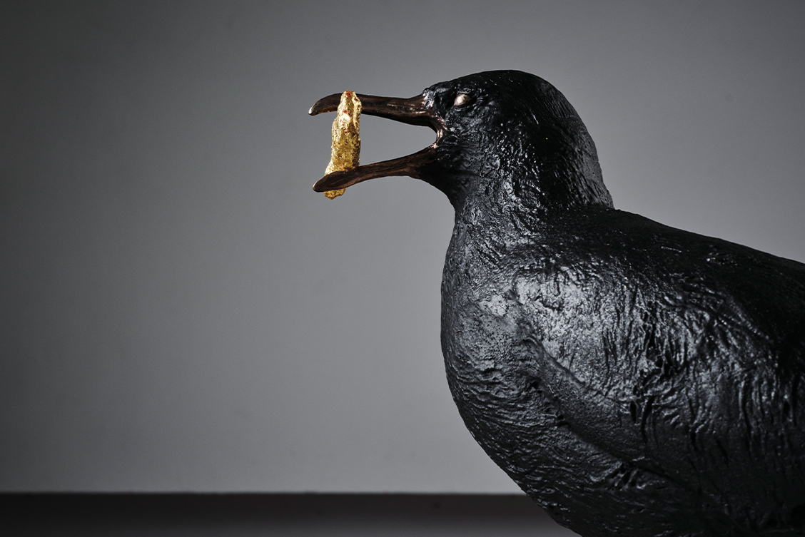 Niamh McCann: someone decides, hawk or dove | Thursday 27 July 2023 |  | Image: photo of a sculpture of a bird, seen side-one, in a plain indoor setting; the bird, which may be a seagull though it is entirely black or bronze-black, is holding something golden in its beak 