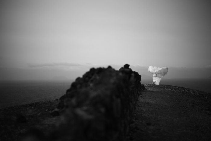 Clare Langan: Songlines 2, 2018. Edition of 3 with 2 artists proofs, 43cm X 28cm. Digital Photograph. Archival Pigment Prints on Hahnemuhle PhotoRag Pearl 320gsm | Clare Langan: Elizium | Saturday 22 July  – Sunday 20 August 2023 | Sarah Walker Gallery | Image: Clare Langan: Songlines 2, 2018. Edition of 3 with 2 artists proofs, 43cm X 28cm. Digital Photograph. Archival Pigment Prints on Hahnemuhle PhotoRag Pearl 320gsm – black-and-white image, apparently taken at the top of a hill; foreground bush / wall (?) blurred, with focus on a figure maybe 100m away, dressed all in white, with the garment billowing upwards and outwards, towards the camera; flat land / sea and clouds in the distance 