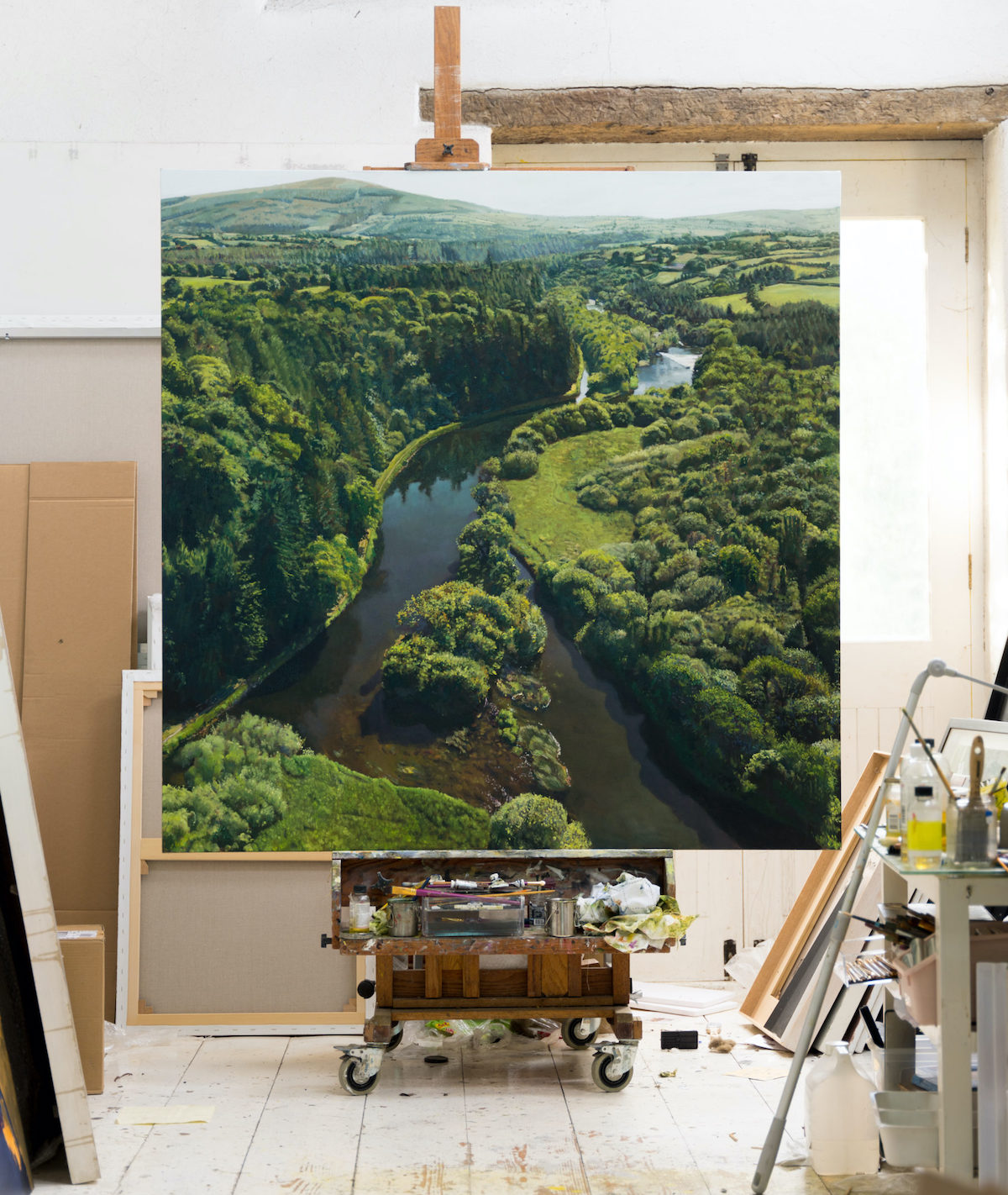 Blaise Smith, Clashganny, oil on canvas, 160 x 160 cm ©Blaise Smith 2023 | Blaise Smith: Irish Landscapes | Friday 8 September  – Sunday 8 October 2023 | Royal Hibernian Academy | Image: Blaise Smith, Clashganny, oil on canvas, 160 x 160 cm © Blaise Smith 2023 – photo taken in an artist’s studio; we see a very large canvas on an easel; the painting – apparently completed – is an aerial view of a multiply forking river in a somewhat hilly landscape of green fields and presumably native trees, all in pale sunshine, probably mid-summer 