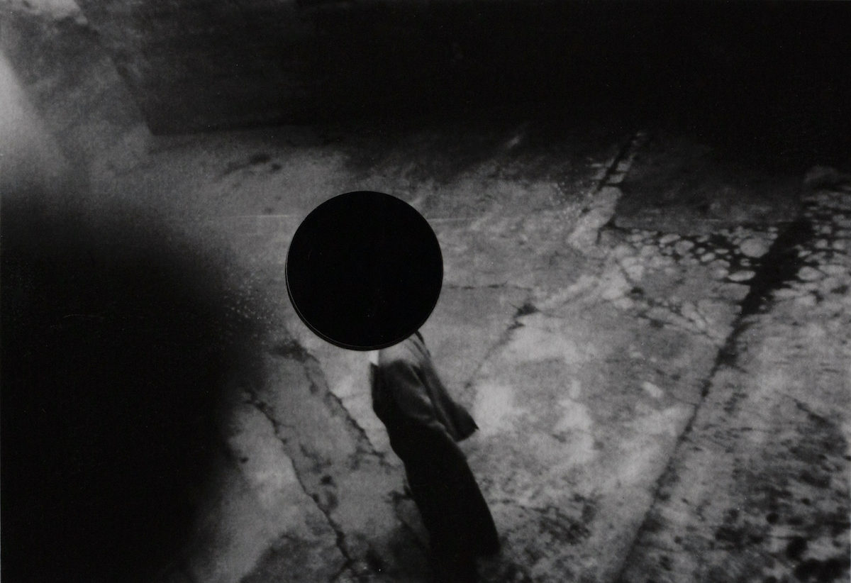 Debbie Castro: Age is a Privilege, Unless You Forget! | Friday 25 August  – Sunday 1 October 2023 | Royal Hibernian Academy | Image: a black-and-white photo of what may be a figure walking in an otherwise empty street scene, viewed from above at an angle; the figure’s head – if it is a figure – is hidden from us by a large black, filled-in circle, a sort of scotoma  