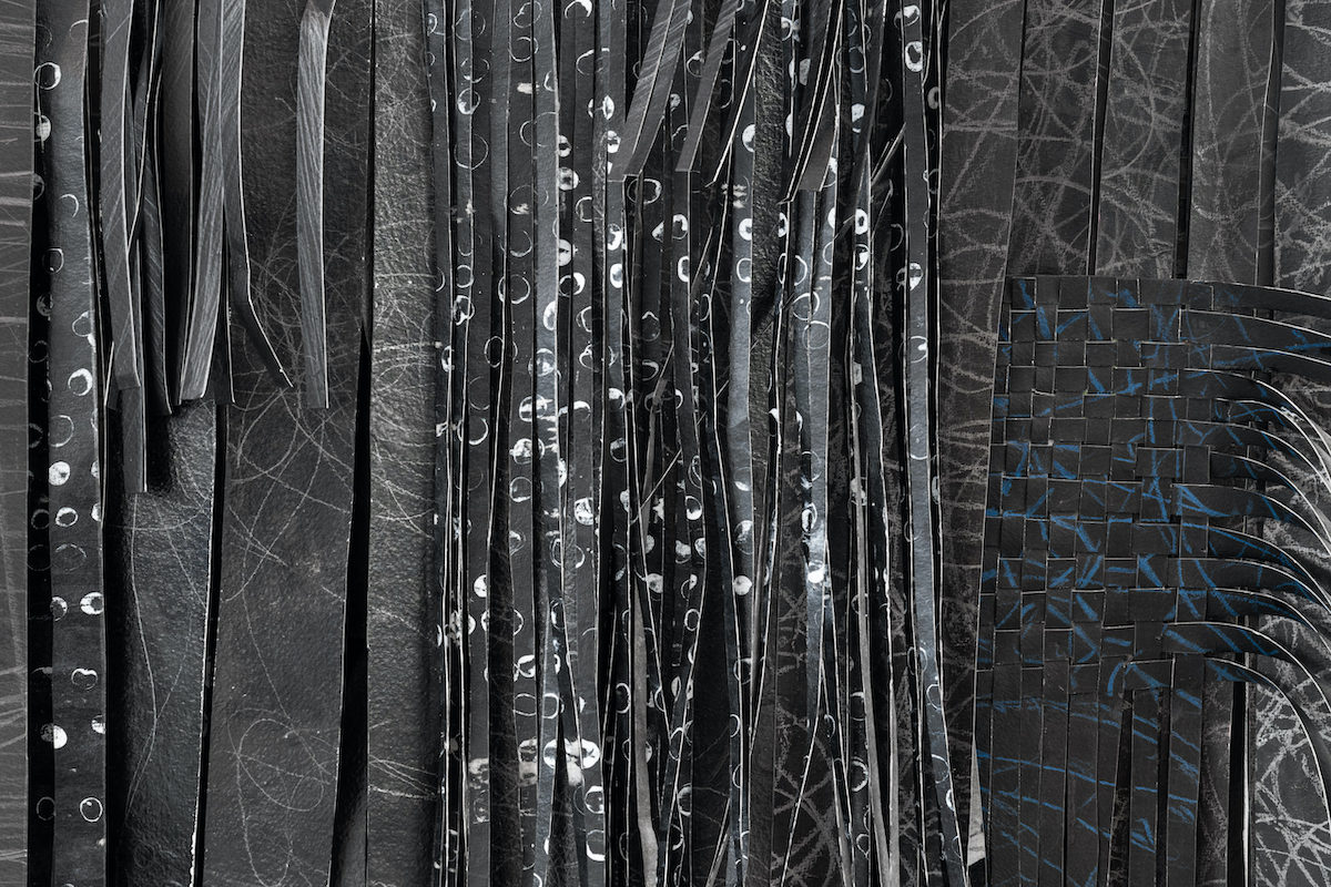 Kiera O’Toole, Weaved Drawing from reworked drawings (detail) 2022, ink, acrylic, graphite on paper, 500 x 500 x 20cm approx. Photography by Jed Niezgoda © | Kiera O’Toole: Weaved Drawing from reworked drawings | Friday 25 August  – Wednesday 1 November 2023 | Royal Hibernian Academy | Image: Kiera O’Toole: Weaved Drawing from reworked drawings, (detail) 2022, Ink, acrylic, graphite on paper, 500 x 500 x 20cm approx. Photography by Jed Niezgoda ©  – it appears that initial artworks have been cut into strips and then these strips have been woven into each other to create a new work; the new work is mostly white on dark grey / black, though some of the marks are blue; most of the strips hang down horizontally, but there is a tighter cross-weaving towards the right, with the horizontal ends of strips bending towards the viewer  