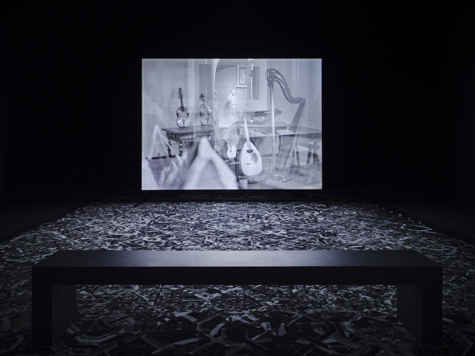 Installation shot Tomasz, M. Inflatable wind Turbine | Bárbara Wagner & Benjamin de Burca: One Hundred Steps | Tuesday 22 August  – Wednesday 4 October 2023 | Wexford Arts Centre | Image: probably an installation shot, in black-and-white, of the film being projected; we see a black simple bench to sit on in the foreground, a richly patterned carpet, then a screen on which is projected a scene showing various stringed musical instruments, possible seen through a window, as there seems to be a reflection that includes a small statue in the distance – or it is a cross-fade 