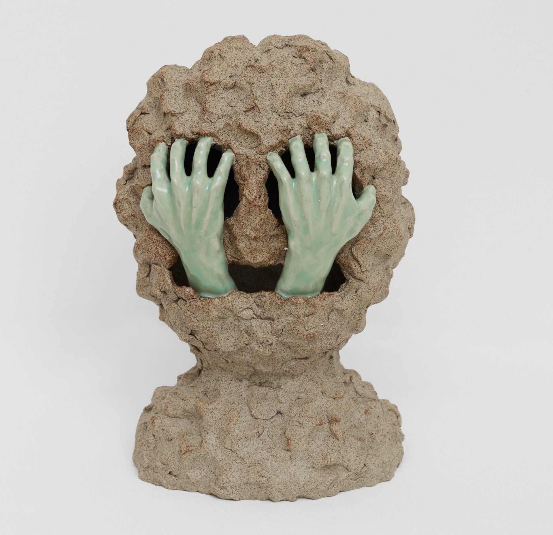 William Cobbing: Grapnel, 2023 | CLAY Festival | Friday 25 August  – Sunday 27 August 2023 | National Sculpture Factory | Image: William Cobbing: Grapnel, 2023 – photo of a clay sculpture; we see a very lumpy round clay head, a bit reminiscent of the Cookie Monster, with large deep holes for the round eyes and a wide horizontal mouth; from the mouth emerges a pair of realistic hands glazed pale green on white; the hands, presumably from the same hidden body, look as though they are clawing at the eyes / eye sockets 