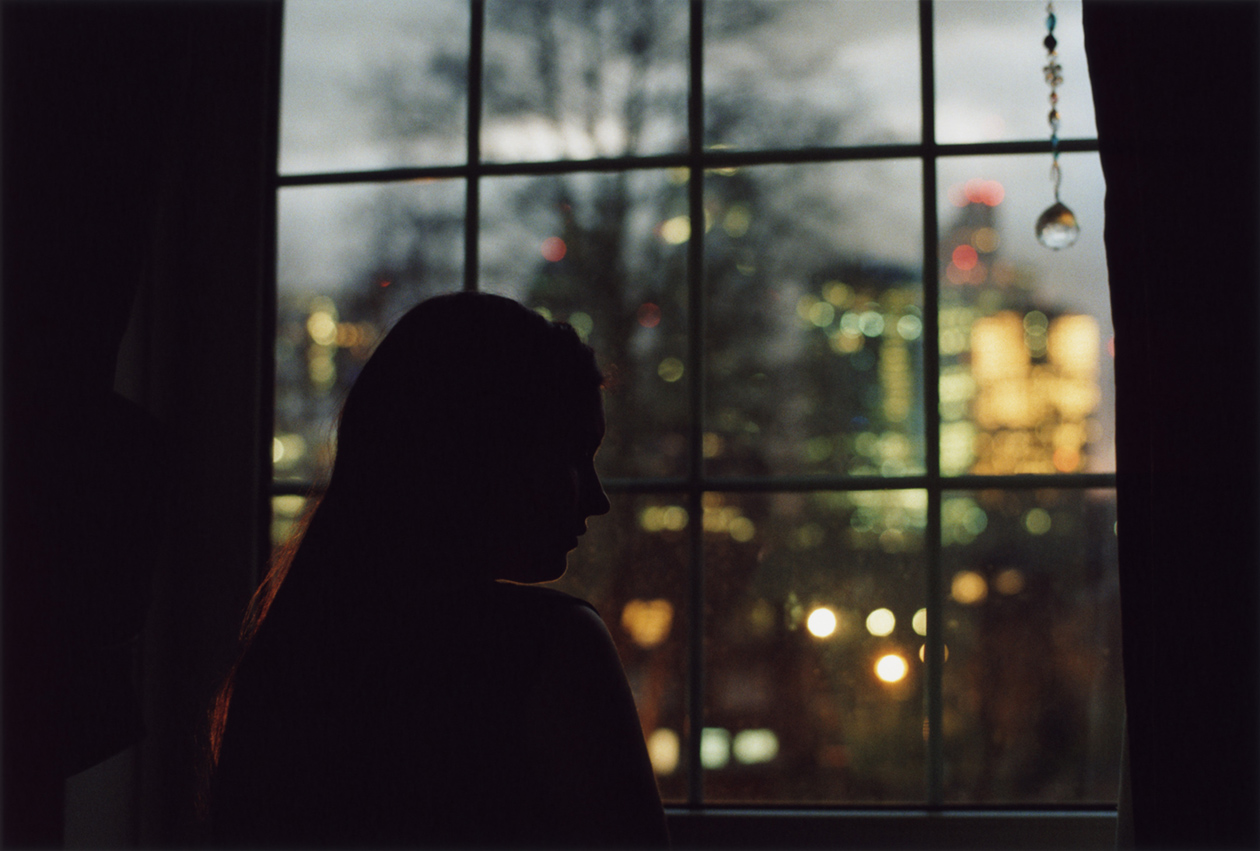 Enda Bowe, from the series Hannah | Enda Bowe: Hannah | Friday 25 August  – Sunday 1 October 2023 | Royal Hibernian Academy | Image: Enda Bowe, from the series Hannah – in the photograph we see the head-and-shoulders dark silhouette of a young woman; the head is in profile, viewed over her right shoulder; she is at a many-paned window through which we have a blurred view of a city at nightfall 