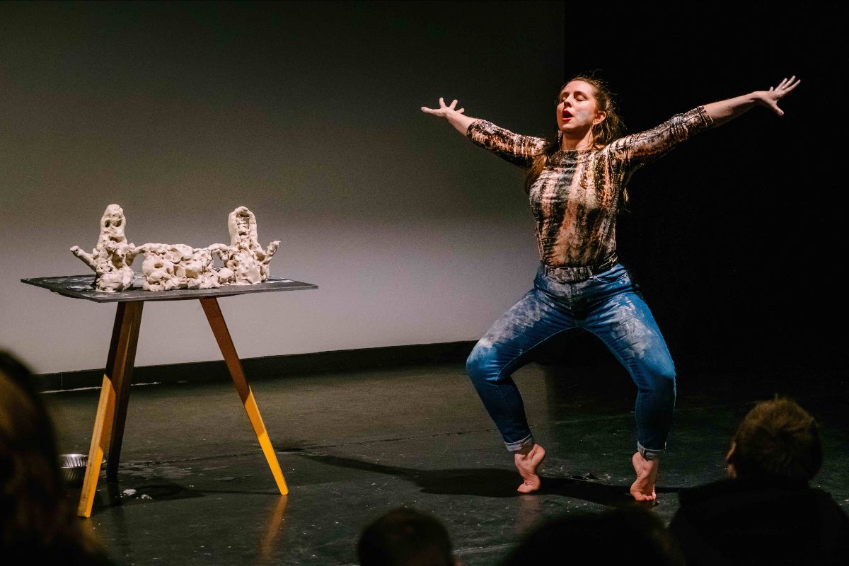 Florence Peake performance, photo Matthew Kaltenborn | CLAY Festival: Florence Peake performance | Sunday 27 August 2023 | National Sculpture Factory | Image: Florence Peake performance, photo Matthew Kaltenborn – photo of the artist during a performance; on the left is a tripod-table with a thin (glass?) top on which are some roughly-modelled clay figures / things; the artist is to the right, on tip-toes and with eyes shut and arms stretched out full-length to the sides and slightly up; the legs are bent, in a sort of quarter-squat; there are clay smudges on the jeans the artist is wearing, suggesting that the clay objects have been worked on during the performance; the scene is strongly lit and we see the backs of heads of some members of the audience 