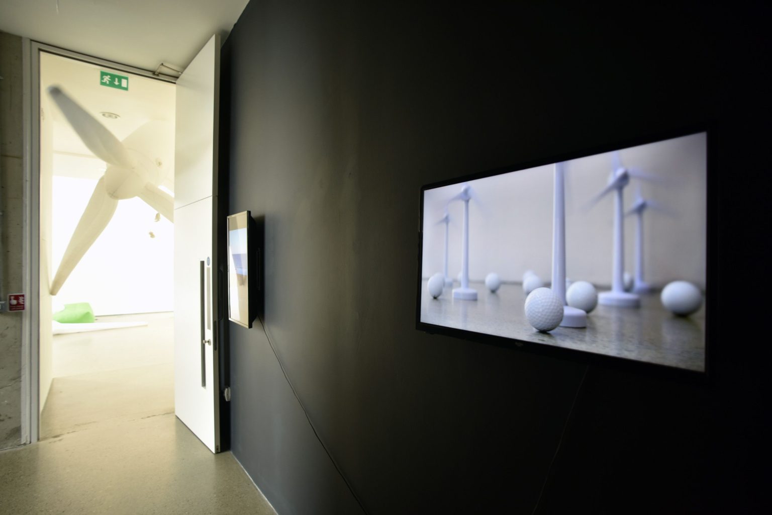 Installation shot Tomasz, M. Inflatable wind Turbine | Micheál O’Connell ¦ Mocksim: System Interference | Tuesday 22 August  – Wednesday 4 October 2023 | Wexford Arts Centre | Image: Installation shot Tomasz, M. Inflatable wind Turbine – a photo possibly taken in a corridor looking into a large room; the black wall of the corridor, taking up about three quarters of the images on the right-hand side, has two images hanging on it; only the nearer is discernible, and it features white wind turbines and white spheres on a reflective surface; these are possibly toys, and the turbines’ blades may be turning; there is a blank, cream-coloured wall behind; in the larger room we glimpse what may be part of an inflatable wind turbine, plus something white and something green in the distance towards a white wall