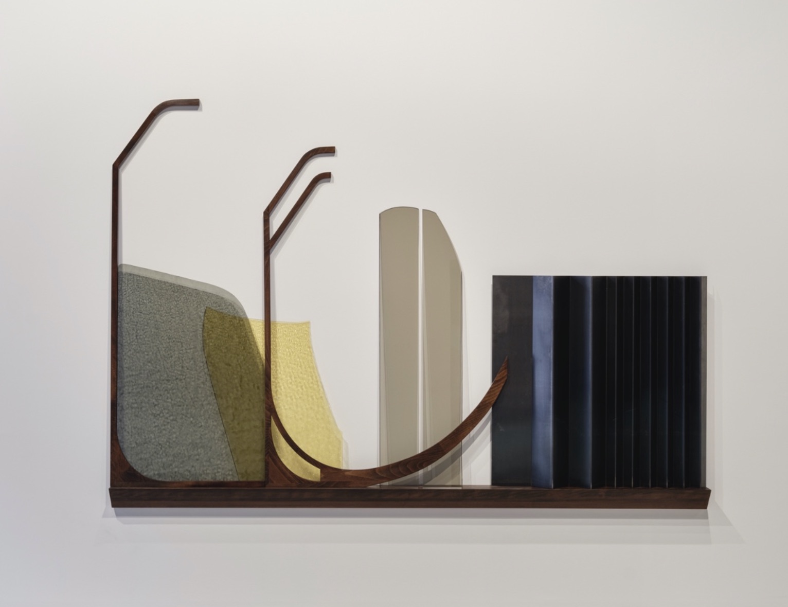 Niamh O’Malley, Shelf, 2022, Beech, glass, steel 1555 x 2245 x 102 mm | Niamh O’Malley: Shelf | Friday 25 August  – Wednesday 1 November 2023 | Royal Hibernian Academy | Image: Niamh O’Malley, Shelf, 2022, beech, glass, steel 1555 x 2245 x 102 mm – photo of various objects on what must be a very long shelf; each object must be almost flat, and each is propped against the white wall on which the shelf is mounted; two or three of the objects – effectively simple shapes – are probably of glass; the other two are of steel or beech; one of them, rust- or wood-coloured, curves in ways that are hard to describe, while the other is square in shape when viewed from the front, but there are hints that it is multiply folded, vertically 