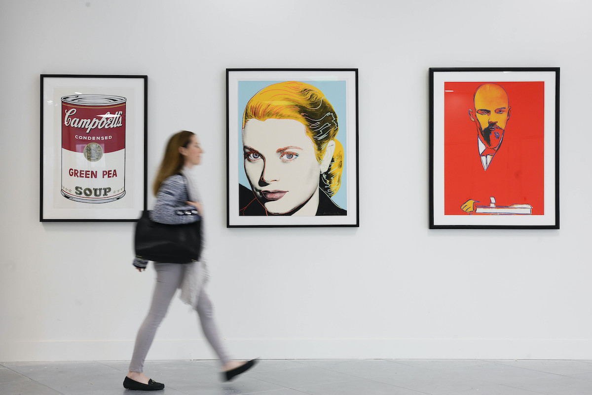 Gormleys at Charlemont Square | Friday 6 October  – Sunday 5 November 2023 |  | Image: photo taken in a gallery space; we see a figure with long hair and a large bag over the right should walking left to right past three prints by Andy Warhol depicting a Campbell’s Soup can, Grace Kelly, and Lenin. 