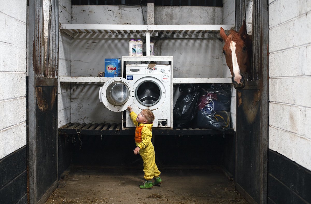 Lorraine Tuck; © the artist | Lorraine Tuck: Unusual Gestures | Saturday 3 February  – Friday 29 March 2024 | Roscommon Arts Centre | Image: Lorraine Tuck; © the artist | the photo is the artist’s young (4?) son, who has ASD, at the door of a washing machine, which is on shelving most of a metre off the ground; he is wearing a yellow costume with an orange front, though we se him from his left side; a horse‘s head is poking in from the right; it is looking at us 
