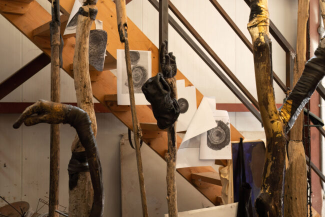 Ben Reilly: Spider Tree | Friday 22 September  – Saturday 18 November 2023 | Garter Lane Arts Centre | Image: photo of various wooden shapes, one of which is also hand-shaped; there are wooden planks in the background, at an angle from bottom right of the photo, more or less, to top left, and pieces of paper – prints? – are attached to these planks / beams | 