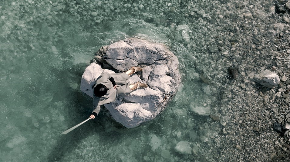 Jasmina Cibic, Beacons (2023), 2 channel film, film still. Courtesy of the artist | Self-Determination: A Global Perspective | Saturday 28 October 2023  – Sunday 21 April 2024 | IMMA | Image: Jasmina Cibic, Beacons (2023), 2 channel film, film still. Courtesy of the artist | film still; view from above of someone sitting on a rock in a river or on a beach; they’ve raised their right high in the air, and it seems to be holding a white stick or baton; they’re wearing a grey overcoat, and we see what looks like some sort of variation on cowboy boots 