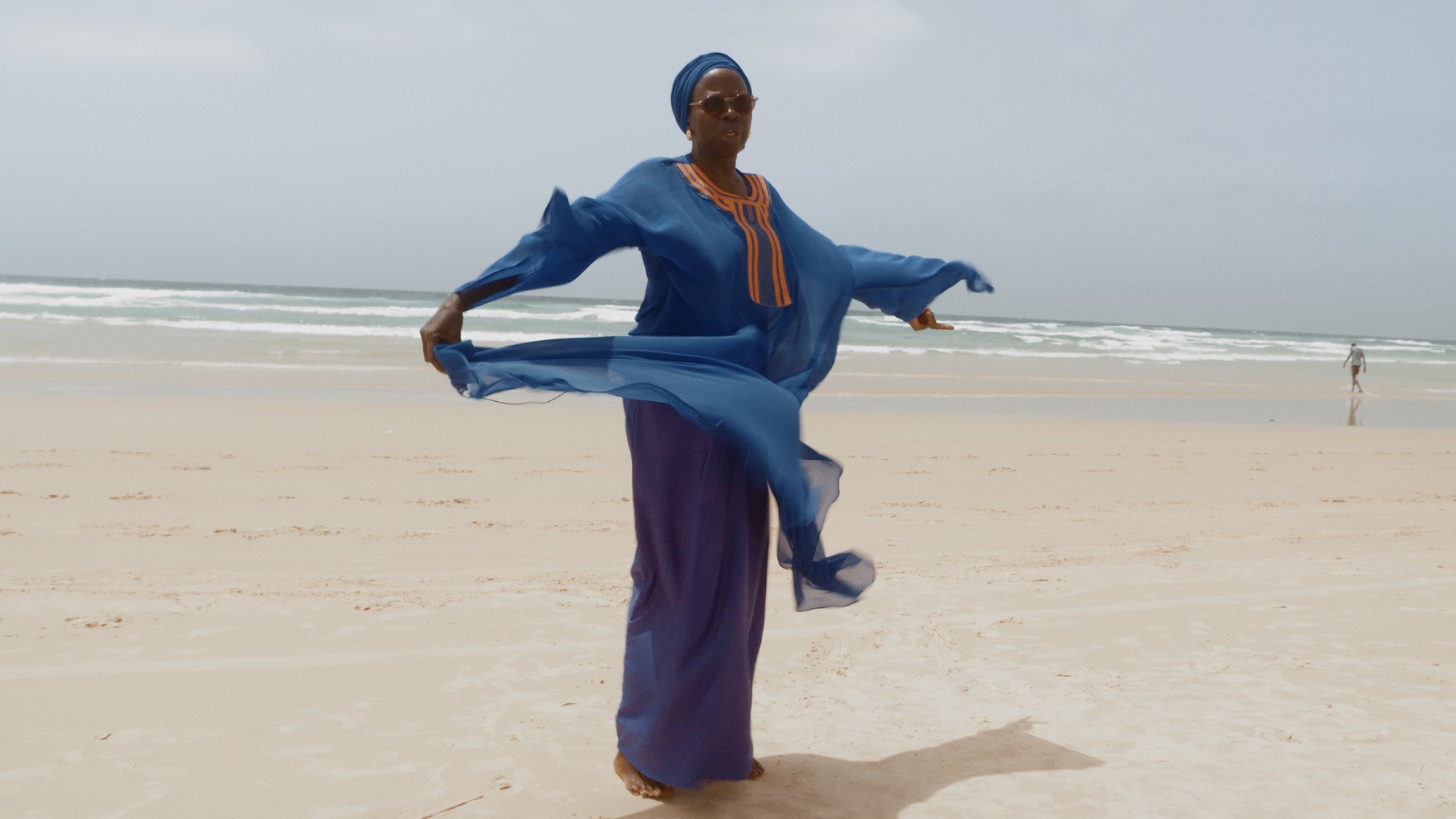 still from Leviathan Cycle Episode 7: Africana, Ken Bugul & Nemo by Shezad Dawood | EARTH RISING | Thursday 21 September  – Sunday 24 September 2023 | IMMA | Image: still from Leviathan Cycle Episode 7: Africana, Ken Bugul & Nemo by Shezad Dawood | someone on a beach in flowing blue, purple and orange clothing, arms outstretched; sunglasses, and face turned towards the camera; beach and sea in the background, stretching to the tilted horizon; figure walking towards the sea towards the right edge of the photo 