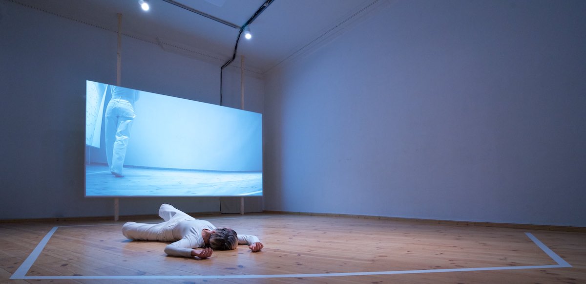 Martijn Tellinga: presence (Un) presence | Thursday 5 October  – Wednesday 11 October 2023 |  | Image: photo of a fairly bare, quite dark space; from a distance of maybe 20 metres, we are looking at one corner of the room; to the left of that corner there is a large screen onto which is projected a photo / video of a figure possibly in the same space; we only see the bottom two-thirds of their body; the video has a strong blue cast; on the wooden floor of the actual space we see a figure clothed in white, on their back, with arms above their head; the head and hands are closest to us | “D-Light Studios is happy to announce Dutch artist Martijn Tellinga’s first solo exhibition in Ireland. After a series of successful EU presentations, ‘presence (Un) presence’ – developed in residence at D-Light – returns to our studios to initiate our new ground floor gallery. With this large-scale, post-industrial space we wish to contribute to Dublin’s vibrant scene and provide an open platform for contemporary and experimental border crossing practices by emerging and established artists…”