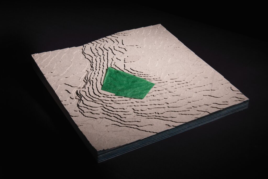 Catriona Gallagher: Sketches for an Artificial Forest | Friday 10 November  – Friday 22 December 2023 | Roscommon Arts Centre | Image: photo – or computer simulation – of a square topographical map viewed from above, corner-on; the map itself is greyish white, with stacked layers of elevation; the map’s square has a certain vertical depth, and the whole is suspended in an entirely black space; on the map a translucent rhomboid has been placed, fairly awkwardly; it may represent the location of a forest 