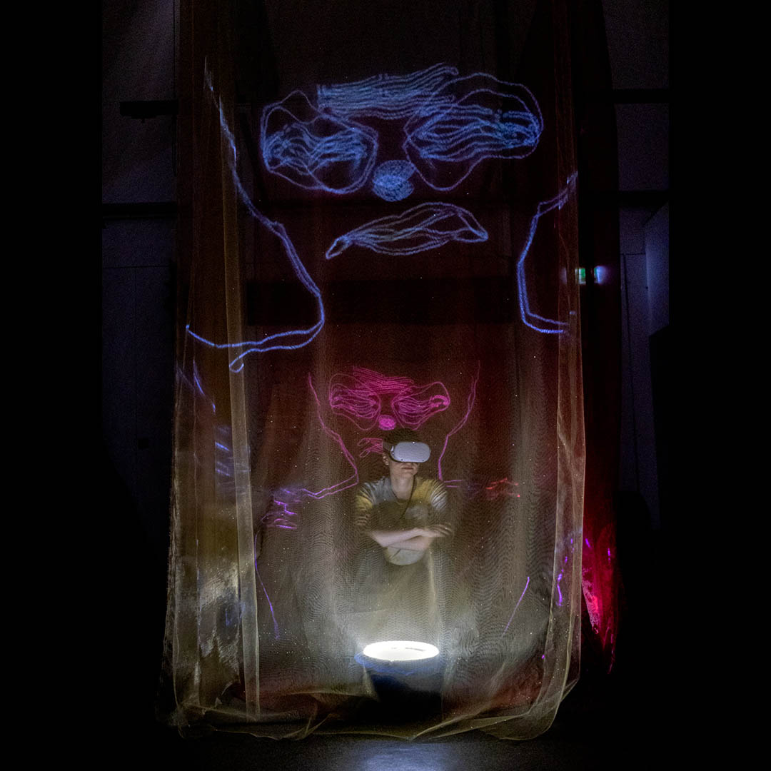 Nikolas Ryan, Transition To Transcendence (2023), VR installation, 2023 | Nikolas Ryan: Transition To Transcendence | Friday 3, Thursday 9 and Friday 10 November  | NCAD Gallery | Nikolas Ryan, Transition To Transcendence (2023), VR installation, 2023 | photo (?) of someone sitting in a fairly spooky environment while wearing white VR goggles; the lighting comes from immediately below, and we see that the space the figure is in is circled by thin gauze curtains; something is projecting, it seems, distorted face outlines onto the gauze in red and pink 