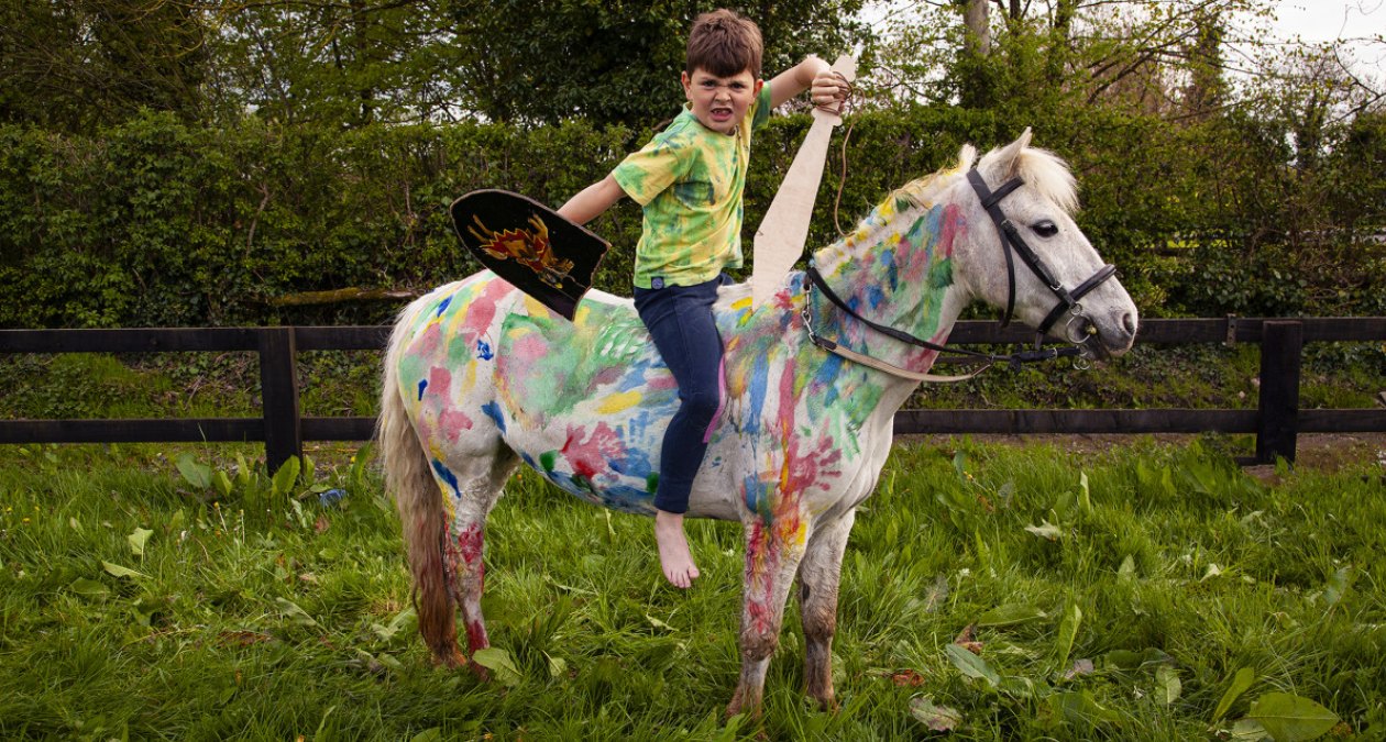 THIS IS ART! | Friday 6 October  – Sunday 29 October 2023 | Royal Hibernian Academy | Image: photo of a kid with toy shield and sword on a white pony in a field; much of teh pony’s coat has been covered handprints and other shapes and splashes of colour; behind the pony and rider are a fence, possibly a narrow road, then a hedge, then trees 
