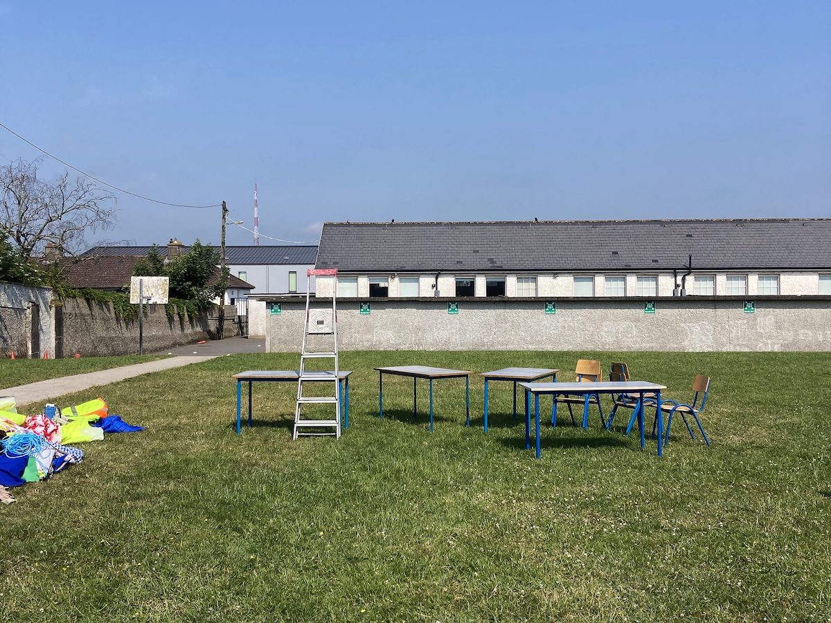 Yvonne McGuinness: Rehearsals | Saturday 21 October 2023  – Sunday 14 January 2024 | Butler Gallery | photo of a large green, grassy area, behind it a long grey, slab-topped wall about 2.5m high, then a long row of windows, white wall and roof; there's a path to the left heading into what looks like a schoolyard; there's the back of the backing board for a low basketball hoop; there’s a wall too stretching from the left, alongside the path but for a metre-wide strip of grass; this wall is part-overgrown and there are two trees rising over it; we see what may be a row of houses beyond the schoolyard, and a very tall, narrow, red-and-white striped chimney in the distance against a somewhat smoggy but otherwise blue sky; in the foreground are four tables, a number of chairs, a ladder leaning against one of the tables, and a pile of colourdy stuff on the left 