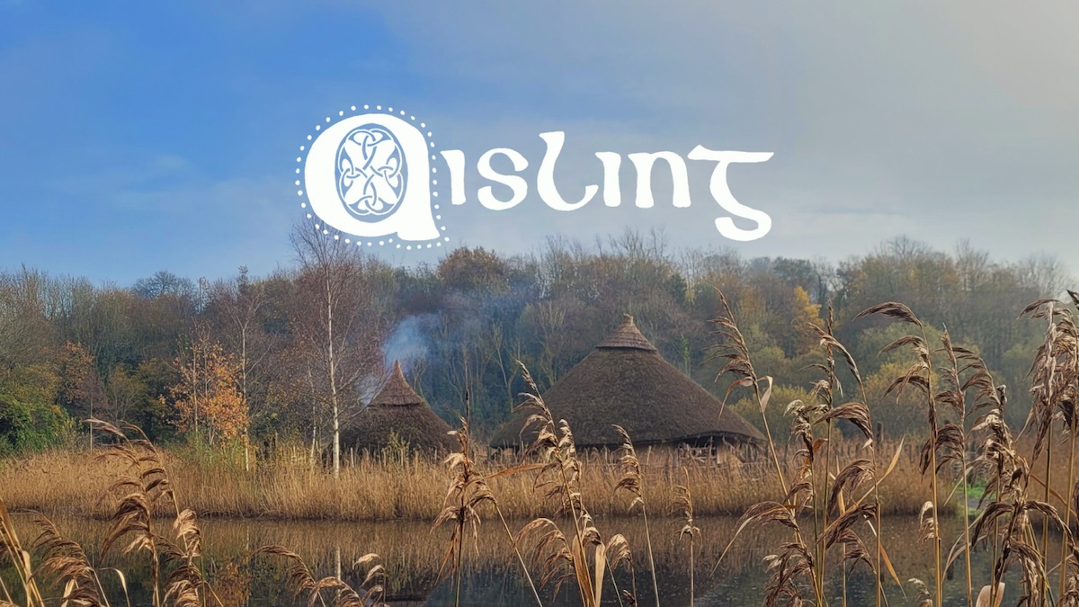 Lorcán Mac Mathúna, Rónán Ó Raghallaigh and John Ó Faoláin: Aisling | Monday 4 December  – Friday 22 December 2023 | Wexford Arts Centre | Image: photo / film still taken in The Irish National Heritage ParkThe Irish National Heritage Park, grasses in the foreground, water, then two reconstructions of prehistorical, circular, thatched dwellings; smoke appears to be rising from one of them; behind is a woods / forest 