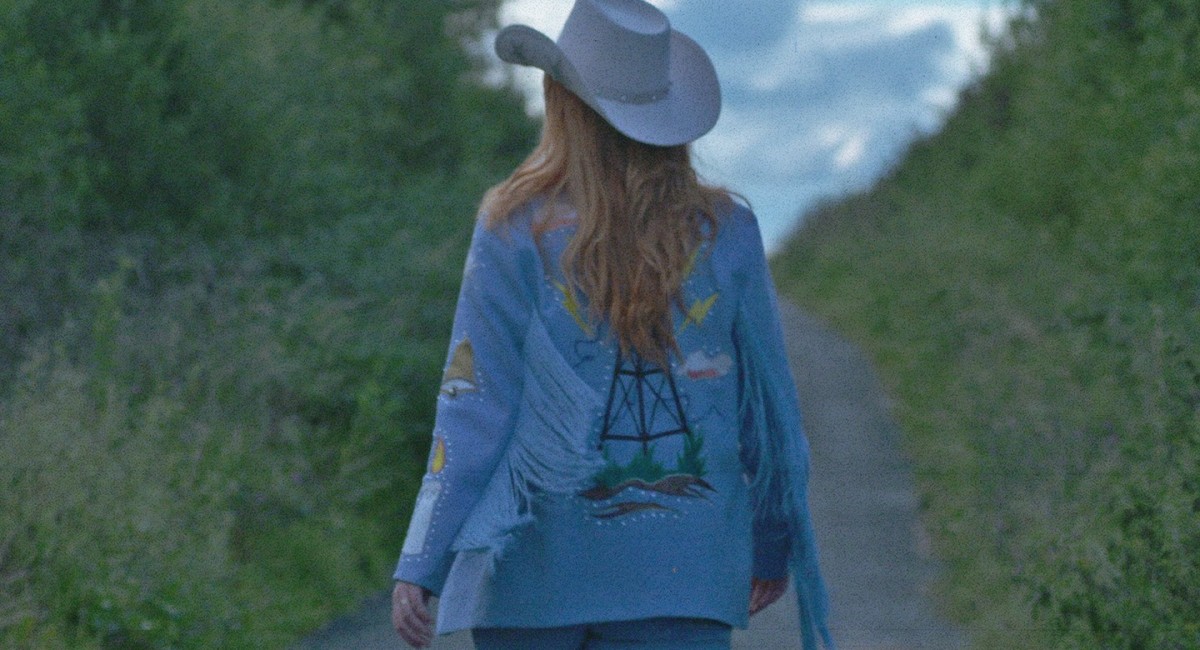 Frank Sweeney: 2 Channel Land still image | Frank Sweeney: 2 Channel Land | Friday 17 November 2023  – Saturday 27 January 2024 | SIRIUS | Frank Sweeney: 2 Channel Land still image | figure in a rural (probably) landscape, seen from behind – stetson hat, long blond hair, blue ‘cowboy’ jacket with various patterns on it, though hard to understand; the figure is apparently on a path sloping upwards between two banks of greenery / high hedges  