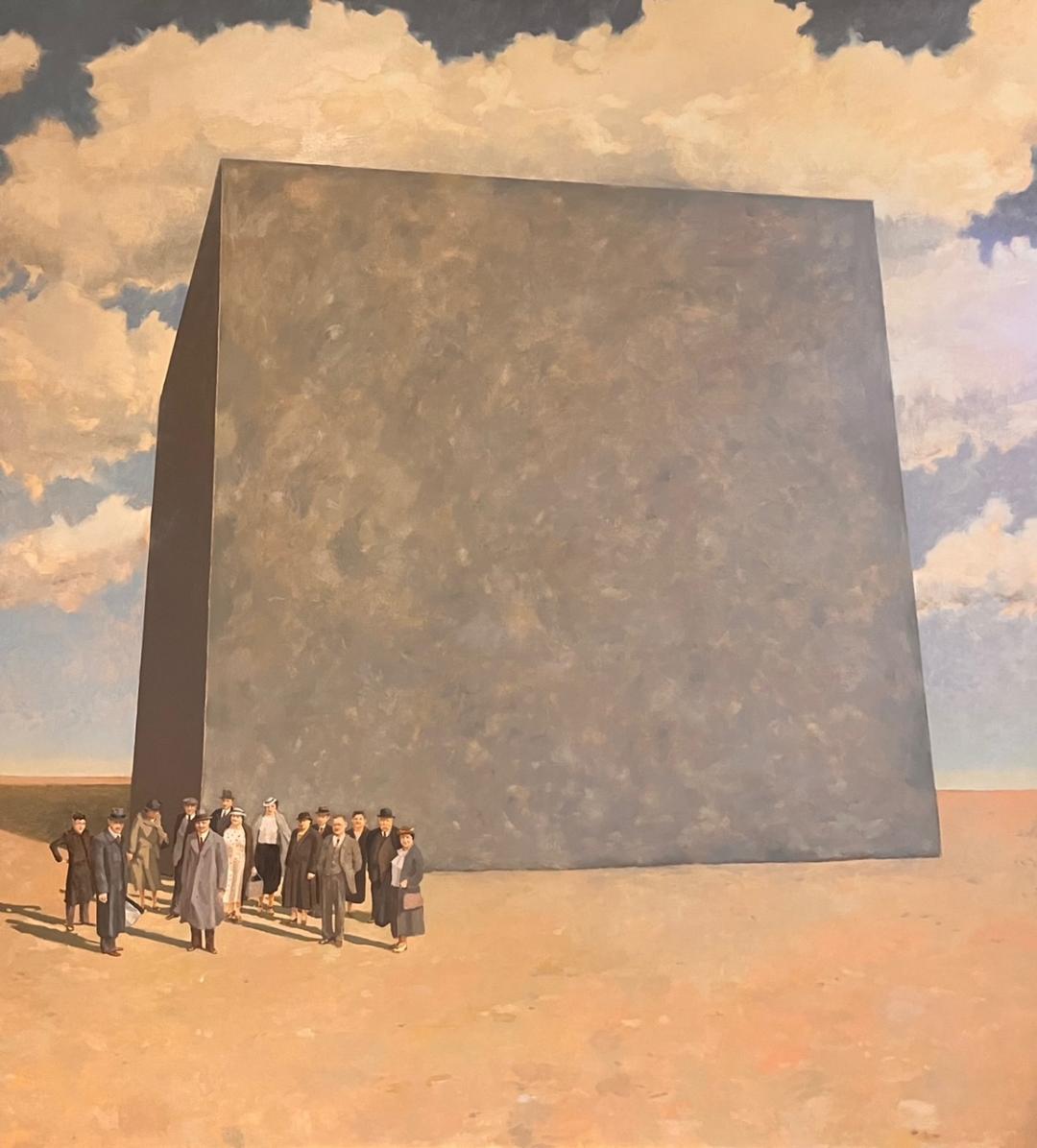 Diarmuid Breen: Bedrock, oil on board, 120 x 136cm | Diarmuid Breen: Give Me My Meaning | Saturday 24 February  – Saturday 18 May 2024 | Garter Lane Arts Centre | Image: Diarmuid Breen: Bedrock, oil on board, 120 x 136cm | In what looks like a flat, empty desert landscape we see a huge grey cube resting like an Egyptian pyramid; it is more or less centred on the canvas and fills most of the canvas; there is blue sky behind, with white clouds, and they may be reflected as well in the surface of the cube; to the left in front of the cube is a group of 14 people dressed as though they might be going to work in an office in some northern city in the 1950s; the attire and the shadows stretching behind them suggest that it could be mid-afternoon in autumn; the style is somewhat reminiscent of Ladybird books 