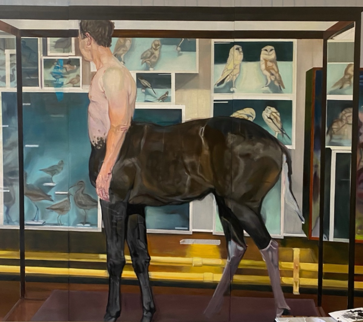 Dominique Crowley, Hibernica Centaurus, Triptych, 2023, Oil and acrylic resin on brace wood panels, 150cm x 240cm, Image courtesy of the artist. | Dominique Crowley: Monstrorum | Thursday 11 January  – Sunday 11 February 2024 | Royal Hibernian Academy | Image: Dominique Crowley, Hibernica Centaurus, Triptych, 2023, Oil and acrylic resin on brace wood panels, 150cm x 240cm, Image courtesy of the artist | painting in a realistic style of a centaur, viewed from the side; the centaur is displayed in a museum vitrine, as in a natural history museum, probably Dublin's ‘dead zoo’; the upper body of the human is male, pale, and pretty ordinary-looking; the head is turned away from the viewer; in the background is a display wall, with images of birds of various sorts; below it we see some yellow-tinted pipes 