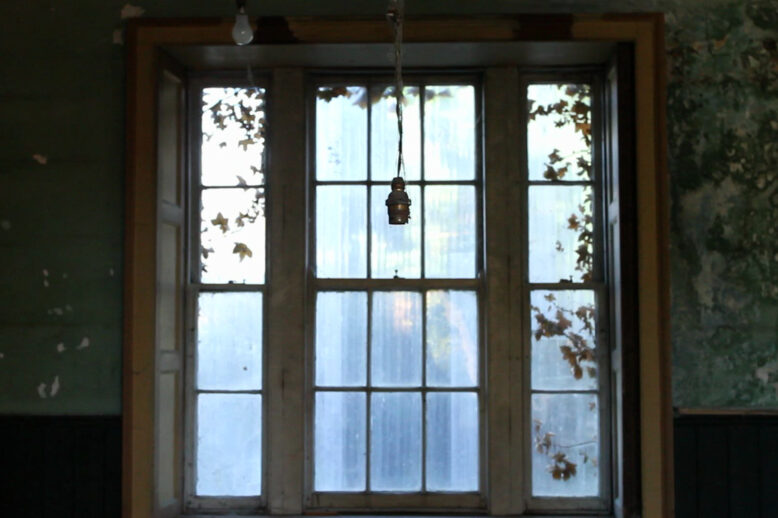 Bridget O’Gorman: : Sometimes the House of the Future is Better Built, 2013, single-channel HD video, surround sound, duration: 6 min 41 sec; camera by Neil O'Driscoll; soundscape by Joe Harney; Butler Gallery Permanent Collection | Bridget O’Gorman: Sometimes the House of the Future is Better Built | Saturday 27 January  – Sunday 24 March 2024 | Butler Gallery | Image: Bridget O’Gorman: Sometimes the House of the Future is Better Built, 2013, single-channel HD video, surround sound, duration: 6 min 41 sec; camera by Neil O'Driscoll; soundscape by Joe Harney; Butler Gallery Permanent Collection | video still of a window seen from inside; it’s in three panels, the two side panels having four panes one above the other, the central panel having treble that amount; the view through the windows is not very clear, possibly due to their being dirty; a lightbulb holder hangs down between us and the window; these are sash windows, and it looks as thought they can be opened nad moved up and down; single-glazed, old, with ivy growing in over the edges outside; we have a partial view of the walls either side, and it looks as though there is a lot of mold or water damage or similar 