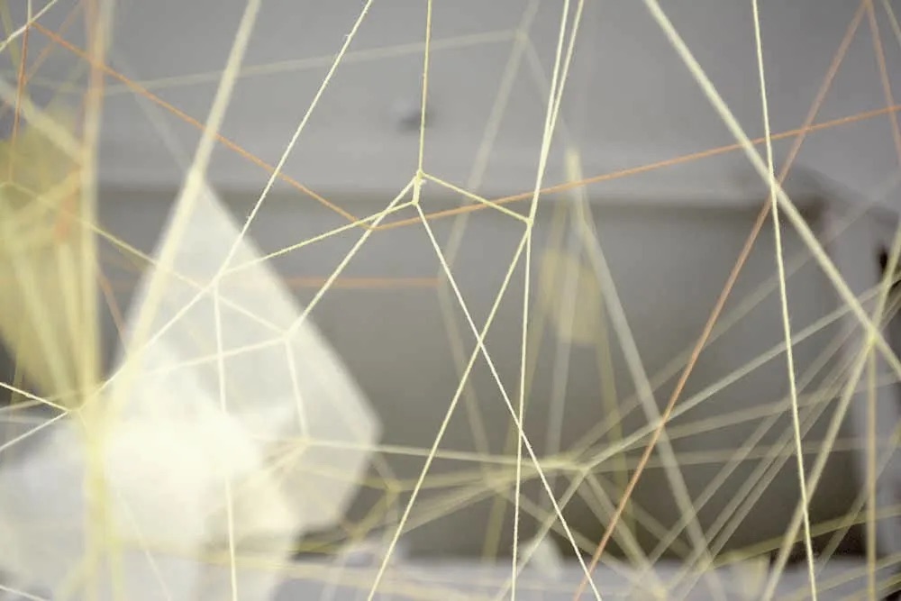 Maia De Faveri, installation shot | Grad Award Exhibition 2024 | Convergence | Thursday 8 February  – Sunday 3 March 2024 | GOMA Gallery of Modern Art | Image: Maia De Faveri, installation shot | photo with plenty of bokeh; criss-crossing yellowish and orange-ish string in the foreground, similar farther back, though with suggestion of shapes of white-ish or yellow fabric or similar material; at the back a wall   