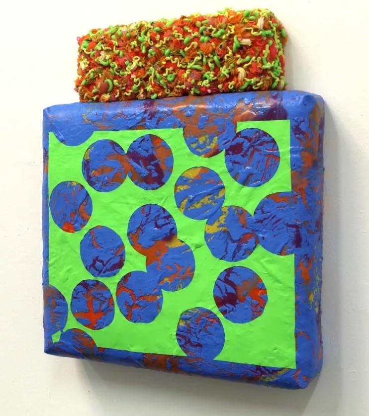 Ciaran Bowen: Sweet Spots, acrylic oil spray paint, silicone paint skins and canvas on expanding foam, 55 x 45 x 9cm | New Horizon | Ciaran Bowen | Wednesday 10 January  – Sunday 4 February 2024 | GOMA Gallery of Modern Art | Image: photo of an artwork mounted on a wall, viewed slightly from one side; it looks like a wrapped present with some sort of decorative sponge on top; the 'present' has a wrapping that is mottled blue, orange and yellow, overlaid with a rectangle of bright green from which random circular shapes, all the same size, have apparently been cut 