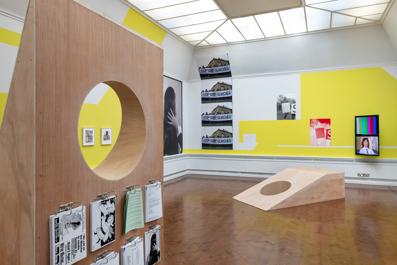 Sara Greavu & Ciara Phillips, We realised the power of it – Derry Film and Video Workshop (2021), 39th EVA International Guest Programme. Courtesy EVA International, Sara Greavu and Ciara Phillips. Photography Jed Niezgoda. | We realised the power of it: Derry Film & Video Workshop | Saturday 30 March  – Sunday 22 September 2024 | IMMA | Image: Sara Greavu & Ciara Phillips, We realised the power of it – Derry Film and Video Workshop (2021), 39th EVA International Guest Programme. Courtesy EVA International, Sara Greavu and Ciara Phillips. Photography Jed Niezgoda. | Installation shot in the Limerick City Gallery of Art, 2021; two plywood constructions with round holes in them; various material on the walls and hanging from one of the plywood constructions; four copies of a 'Stop Strip Searches' protest photo, in a vertical column towards back left of the room 