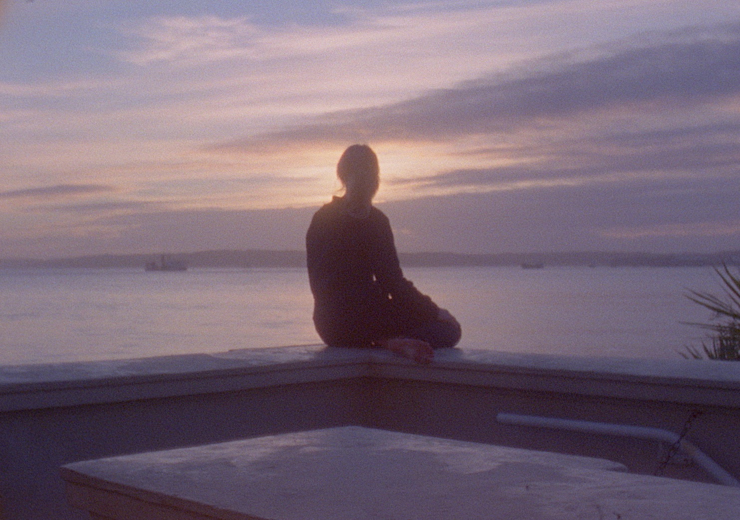 Aoife Desmond, sirius – harbour’s mouth (still), 2023. Courtesy of the artist | Aoife Desmond: sirius – harbour’s mouth | Saturday 17 February  – Saturday 16 March 2024 | SIRIUS | Image: Aoife Desmond, sirius – harbour’s mouth (still), 2023. Courtesy of the artist | we see a figure sitting on a parapet and looking out to sea – or more likely to Cork harbour; the figure’s back is to us, there is a suggestion of long hair, and we see the sole of the figure’s bare right foot; there are two or more ships in the bay; it is probably evening, with the figure’s head blocking the sun, which is fairly low; there’s a warmish, greyish tint to everything 