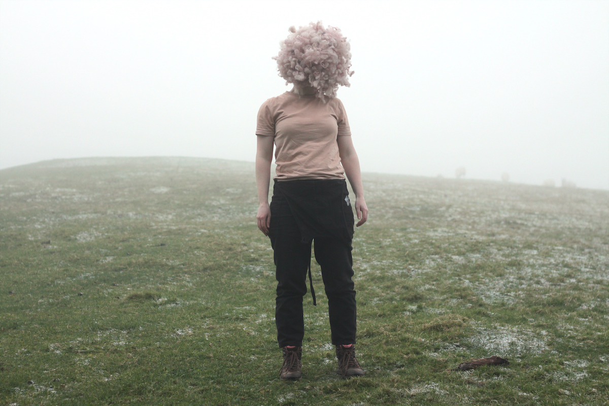 Joanna Hopkins: Fruity Bodies | Saturday 9 March  – Friday 3 May 2024 | Municipal Gallery | Image: photo of female standing in a misty field; wearing black jeans and a pink T shirt, with a fluffball where the head should be 