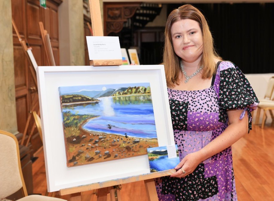 Niamh Eldridge-Barry | Connections Arts Centre at the RHA | Friday 16 February  – Sunday 28 April 2024 | Royal Hibernian Academy | Image: Niamh Eldridge-Barry | The artist is displaying a framed painting on an easel; the painting is a realistic depiction of a what is probably a river scene on a bright day; there are strong shadows of rocks on flat red soil in the foreground; hills stacking towards the horizon, the nearest treed; the sun is level with the top of the hills, and it leaves a band of bright light in the water, stretching towards the viewer  