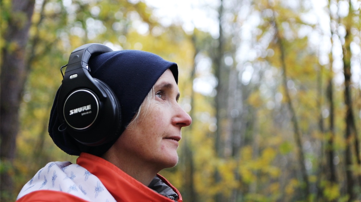 Karen Power: wonderous worlds within / without: interspecies gossip | Saturday 2 March  – Saturday 27 April 2024 | SIRIUS | Image: Karen Power. Photograph: Frida Sjögren | almost-profile photo of the composer, who is looking from left to right; just the head and top of right shoulder; dark-blue woolly hat with headphones over them; blurred background of autumnal (probably) trees 