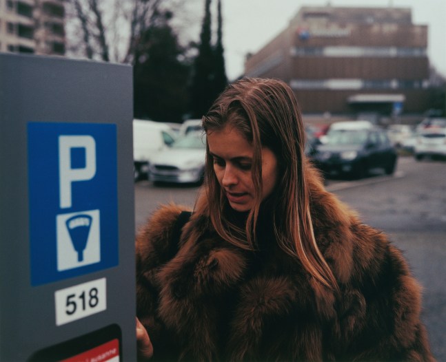 Samuel Laurence Cunnane: Fur, 2023, hand-printed C-type print on archival photo paper, framed, edition of 3 + 1AP, 32 x 39.5 cm / 12.6 x 15.6 in image size | Samuel Laurence Cunnane: Late Spring | Friday 1 March  – Saturday 6 April 2024 | Kerlin Gallery | Image: Samuel Laurence Cunnane: Fur, 2023, hand-printed C-type print on archival photo paper, framed, edition of 3 + 1AP, 32 x 39.5 cm / 12.6 x 15.6 in image size | photograph; we see head and shoulders of a long-haired brunette figure, wearing a very furry fur coat, at a parking-payment machine; the face is looking down, to where a just-visible hand is interacting with the machine; behind is an unremarkable open carpark with a large dull-red building behind; blurred trees suggest that it is early spring  