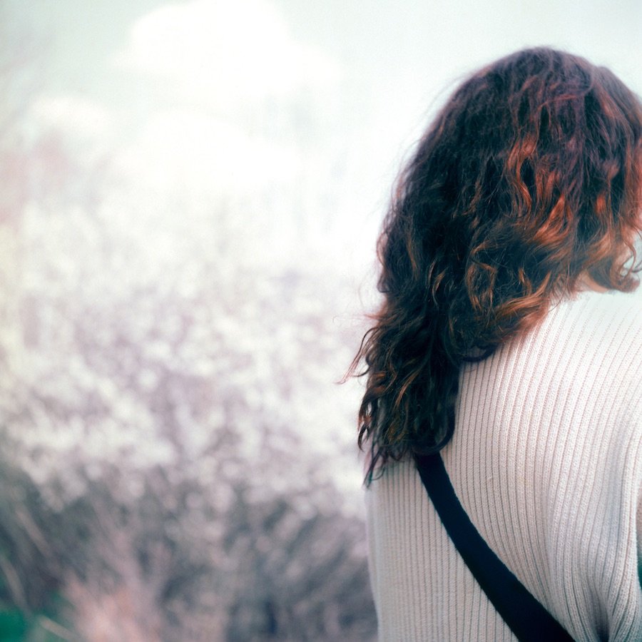 Leon Nevill Gallagher, Untitled, from the series Cargo | Imprints: Photography as Practice | Thursday 22 February  – Saturday 16 March 2024 | Photo Museum Ireland | Image: Leon Nevill Gallagher, Untitled, from the series Cargo | this is a somewhat hazy photo, with quite a blurry background that suggests a bush of some type which stretches upwards towards a sort of mushroom cloud, which seems unlikely…; there is a figure to the right, taking up about a third of the photo and partially cropped by the edge of the photo; we see long brown-to-black partially sunlit hair coming down over a light, white, ribbed jumpber; there is also a black band as of an unseen bag crossing over the back of the subject, whose face we do not see 