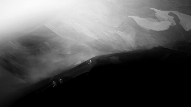 Clare Langan: The Heart of a Tree, film still (2020), Collection Crawford Art Gallery, Cork. Image © the Artist | Clare Langan: The Heart of A Tree  | Thursday 14 March  – Sunday 28 April 2024 | Crawford Art Gallery | Image: Clare Langan: The Heart of a Tree, film still (2020), Collection Crawford Art Gallery, Cork. Image © the Artist // black-and-white photo; we see three figures in the distance in what looks like a snowy, volcanic landscape, with added mist