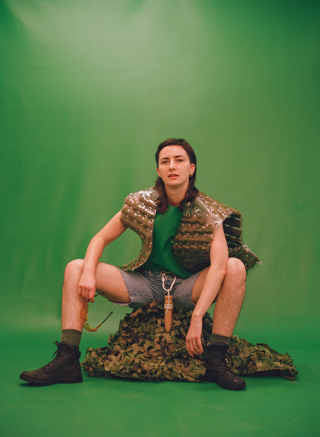 Beyond Survival Expert (2022). 120mm photograph, photographer: Niamh Barry, assistant photographer: Aoife McGrath, image courtesy of Léann Herlihy and Project Arts Centre | Apocalypse Anxieties | Saturday 27 April  – Thursday 27 June 2024 | Luan Gallery | Image: Beyond Survival Expert (2022). 120mm photograph, photographer: Niamh Barry, assistant photographer: Aoife McGrath, image courtesy of Léann Herlihy and Project Arts Centre | figure in a photo studio, long, straight dark har to below shoulders; looking straight at us, sitting on what looks like a mound of camouflage gear; wearing what looks a sort-of armoured gilet, green t-shirt, grey shorts, green socks, walking boots; wooden dowel hanging down between legs 