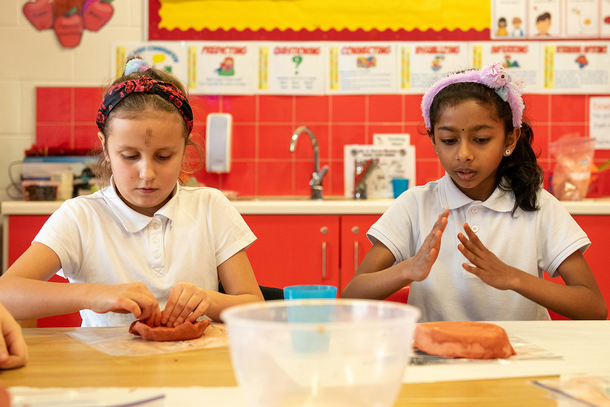Living Arts Project 2024 | Saturday 30 March  – Wednesday 1 May 2024 | Wexford Arts Centre | Image: two schoolkids seated at a table, modelling clay; we’re looking from across the table; they have white to[ps and colourful headbands; behind them is what looks somewhat like a kitchen sink, with a white counter top, red cupboard and red tiles on the wall; we’re presumably in the craft room of the school; the schoolkid on the left is modelling clay with both hands, the one on the right is gesturing, both hands near each other and floating above what seems to be a large, wet, round plop of modelling clay 