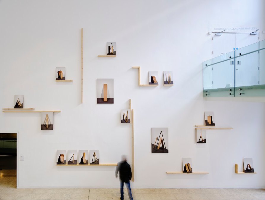 Liam O’Callaghan: From the Mass Presence of Possibilities…, exhibition installation shot | Liam O’Callaghan: From the Mass Presence of Possibilities… | Friday 16 February  – Sunday 28 April 2024 | Royal Hibernian Academy | Image: Liam O’Callaghan: From the Mass Presence of Possibilities…, exhibition installation shot | photo taken in the inner foyer / stairwell of the RHA; 12 paintings are distributed on shelves at various heights and left-right positions across a large, high, white wall, and a further five appear to be hung, rather than on shelves; the paintings depict pieces of wood / board leaning against walls  
