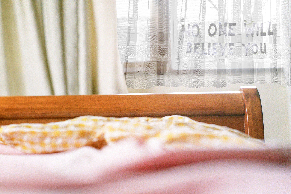 Suella Holland: No One Will Believe You (detail), medium format film photograph, 16 x 16 inches | Suella Holland: A Joining of Self | Saturday 13 April  – Saturday 8 June 2024 | Solstice Arts Centre | Image: Suella Holland: No One Will Believe You (detail), medium format film photograph, 16 x 16 inches | photo taken apparently in a bed, probably looking towards the foot of the bed, marked by a wooden bed-end, then there appears to be a gap, then a window with cream curtain and lace / gauze curtain; the latter seems to have the words ‘NO ONE WILL BELIEVE YOU’ woven into it or attached to it; outside there's a hint of sky and maybe a tree branch  