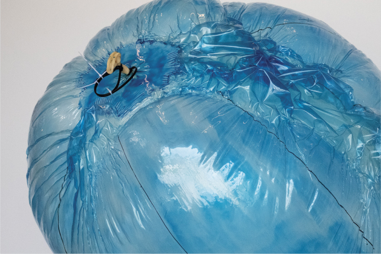 Helen Hughes: Untitled, 2024. 90 x 80 x 90 cm, balloon, rubber, plastic ties (detail) | Helen Hughes: finding the most forgiving element | Saturday 6 April  – Sunday 26 May 2024 | Butler Gallery | Helen Hughes: Untitled, 2024. 90 x 80 x 90 cm, balloon, rubber, plastic ties (detail) | photo of the top of a blue-ish, translucent balloon; there’s a bit of khaki plastic sticking out of it and what may be a black rubber tie; the balloon itself is not of the normal party type; instead it appears to be joined-together strips of balloon material; the joins are visible, and the balloon surface curves inwards towards the joins; the background is a slightly pink grey 