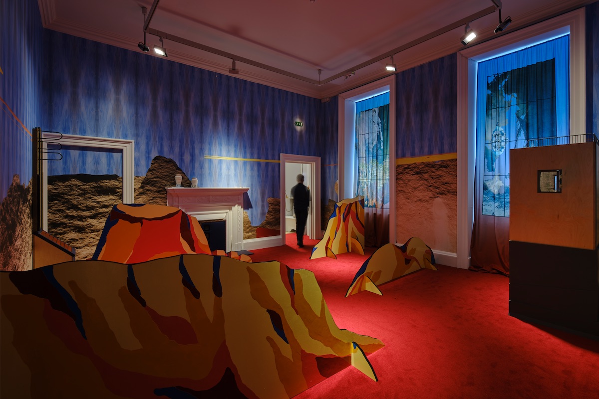 Array Collective. Installation view Self-Determination: A Global Perspective, IMMA, Dublin 2024 / Photo Ros Kavanagh. | Self-Determination: A Global Perspective – Artists Commissions |   to Sunday 15 September 2024 | IMMA | Image: Array Collective. Installation view Self-Determination: A Global Perspective, IMMA, Dublin 2024 / Photo Ros Kavanagh | photo of a room at IMMA; funky blue patterned wallpaper, sort of neuronal-looking;  unpatterned red carpet throughout; white Georgian-style fireplace, with two white vases (?) on it; large white frame to the left in the room, framing nothing; part of the wallpaper is hidden / papered over by what looks roughly like a pattern of mud or dirt, but dry; silly fake cardboard or hardboard mountains, flat, held up by 'feet' of the same material; pattern of ridges on them, from a dry, hot country; blur of someone passing through tghe doorway at the back; other strange bits and pieces; museum lighting from the ceiling 