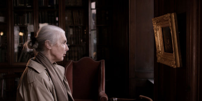 © Martin Healy, 2016 | Martin Healy: A Moment Twice Lived | Thursday 2 May  – Sunday 30 June 2024 | Crawford Art Gallery | Image © Martin Healy, 2016 | photograph of a figure in what appears to be a dark, fancy living-room or something similar; we see a figure towards the left, in profile and looking across to the right, where we see a framed artwork (the frame is fancy, gold, the artwork A4 or smaller, dark, seen side-on more or less; the figure on the left has greying hair, shoulder-length but tied up by a ribbon at the back; pale-brown scarf and slightly paler raincoat; we see down only as far as the upper arm 