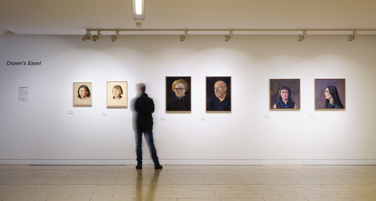 Orpen’s Easel | Saturday 17 February  – Sunday 28 April 2024 | Royal Hibernian Academy | Image: photo taken in the entrance area of the RHA; there are six portrait paintings hanging on the wall, in pairs by the artist who painted them; to the left of the artowrks is the shows title, on the wall, and an explanatory text; a black-clad, motion-blurred figure is standing between the first and second pairs of paintings 